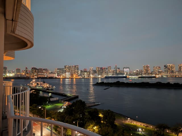 A cityscape at dusk with illuminated buildings across a body of water, viewed from a balcony