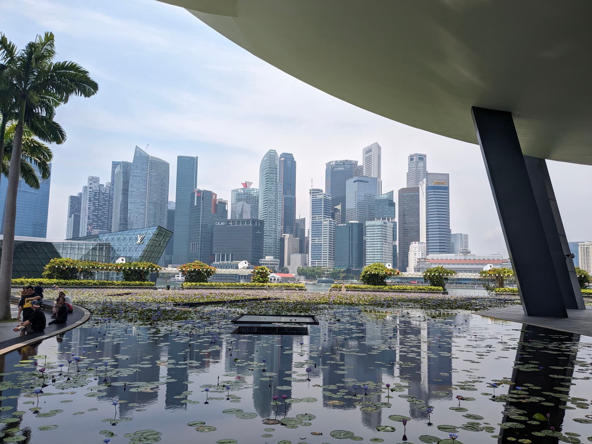 A city skyline with modern skyscrapers is seen across a reflective water feature, framed by architectural elements and palm trees