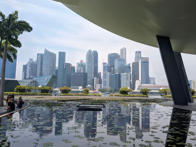A city skyline with modern skyscrapers is seen across a reflective water feature, framed by architectural elements and palm trees