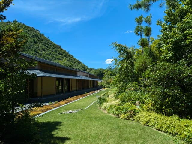 A serene landscape featuring a traditional building surrounded by lush greenery and a manicured lawn, set against a backdrop of a forested hill under a clear blue sky